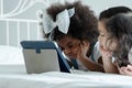 Selective focus on little African kid girl with bow tie on afro hair is lying on white bed put hands on chin and using tablet Royalty Free Stock Photo