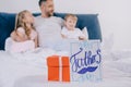 Focus of red gift box and happy fathers day greeting card near cheerful man embracing son and daughter while sitting on Royalty Free Stock Photo