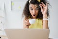 Focus of shocked african american girl touching glasses while looking at laptop and holding cup of coffee Royalty Free Stock Photo