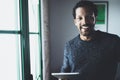 Selective focus.Smiling bearded African man looking at camera and holding in hand digital tablet near the window in