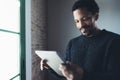 Selective focus.Smiling bearded African man reading news digital tablet while standing near the window in his modern Royalty Free Stock Photo