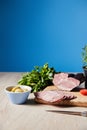 Focus of tasty ham on cutting board with knife, parsley, olives on wooden table on blue background