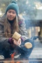 Teenage girl wearing beanie hat roasting large marshmallow on a stick over the campfire firepit. Camping family fun lifestyle