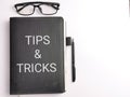 Selective focus top view text TIPS AND TRICKS on black book with pen and eye glasses isolated on white background. Royalty Free Stock Photo