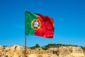 Selective focus on waving flag of portugal in front of typical yellow rock formation of algarve and blue sky. National portugal