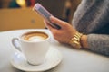 Close-up of white cup of coffee on the table in a modern cafe and woman hand with clock holding smartphone. Royalty Free Stock Photo