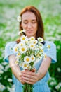 Selective focus a young woman in a blue dress holds a bouquet of daisies in front of her on the background of a chamomile field