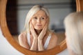 Self-care concept. Beautiful senior woman looking at mirror, touching soft skin on face and enjoying her reflection