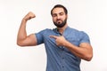 Self confident egotistical bearded man in blue shirt pointing finger at his arm biceps, proud and satisfied with muscular build,