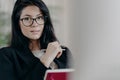 Self confident serious adult female employee looks though transparent glasses, writes information while working on project, makes