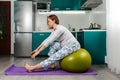 Self-isolation and home training. A pregnant Caucasian woman sits on a fitball, and makes bends to the feet. Kitchen in the