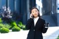 Self-possessed asian girl talking on the phone, business woman near the office center on a break, holding a phone and a coffee