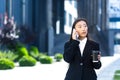 Self-possessed asian girl talking on the phone, business woman near the office center on a break, holding a phone and a cup of hot
