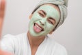 Selfie Glam: lady in bathrobe, snaps a selfie with a clay mask, showing tongue at the camera.