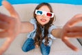 Selfie portrait from above of amazing joyful little girl in 3d glasses, jeans clothes expressing to camera on couch on