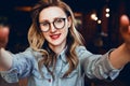 Selfie portrait of young smiling woman sitting in cafe. Hipster girl in trendy glasses takes a selfie in coffee shop.