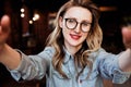 Selfie portrait of young smiling woman sitting in cafe. Hipster girl in trendy glasses takes a selfie in coffee shop. Royalty Free Stock Photo