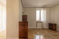 A semi-empty room with some vintage-style wooden furniture with a window with curtains and an exit to a walk-in hallway with