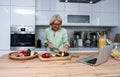 Senior adult woman cooking healthy food for dinner in her domestic kitchen using laptop computer to read online recipe. Royalty Free Stock Photo