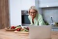 Senior adult woman cooking healthy food for dinner in her domestic kitchen using laptop computer to read online recipe. Royalty Free Stock Photo