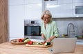 Senior adult woman cooking healthy food for dinner in her domestic kitchen using laptop computer to read online recipe. Royalty Free Stock Photo