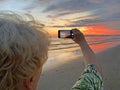 Senior adventure is ageless woman with a mobile phone take a photo of beautiful of sunset near sea beach. Summer vacation for Royalty Free Stock Photo