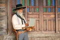 Senior Asian cowboy stand in front of wooden building and use laptop alone in concept of technology for senior people in daily