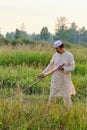 An senior asian man in an embroidered skullcap and white traditional clothes mows hand-scythe grass in a hayfield