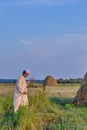 An senior asian man in an embroidered skullcap and white traditional Clothes mows hand-scythe grass in a hayfield