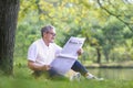 Senior Asian man reading a business newspaper while sitting under the tree by the lake at the public park for recreation, leisure Royalty Free Stock Photo