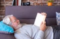 Senior attractive man lying down on sofa reading a book.  Old retired man relaxing at home.  Brick wall background Royalty Free Stock Photo