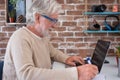 Senior attractive man using laptop computer at home smiling. Brick`s wall on background