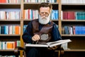 Senior bearded man in glasses, academic professor or teacher, sitting and reading an old book in the library, holding Royalty Free Stock Photo