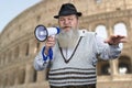 Senior bearded man talking into megaphone while standing on blurred background of Colosseum at Rome, Italy.