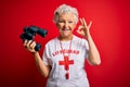 Senior beautiful grey-haired lifeguard woman using binoculars and whistle over red background doing ok sign with fingers,