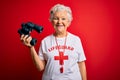 Senior beautiful grey-haired lifeguard woman using binoculars and whistle over red background with a happy face standing and