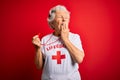 Senior beautiful grey-haired lifeguard woman wearing t-shirt with red cross using whistle bored yawning tired covering mouth with