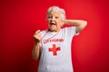Senior beautiful grey-haired lifeguard woman wearing t-shirt with red cross using whistle Crazy and scared with hands on head,