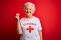 Senior beautiful grey-haired lifeguard woman wearing t-shirt with red cross using whistle with a happy and cool smile on face