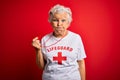 Senior beautiful grey-haired lifeguard woman wearing t-shirt with red cross using whistle puffing cheeks with funny face