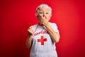 Senior beautiful grey-haired lifeguard woman wearing t-shirt with red cross using whistle shocked covering mouth with hands for