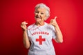 Senior beautiful grey-haired lifeguard woman wearing t-shirt with red cross using whistle smiling doing phone gesture with hand