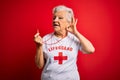 Senior beautiful grey-haired lifeguard woman wearing t-shirt with red cross using whistle smiling with hand over ear listening an