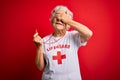 Senior beautiful grey-haired lifeguard woman wearing t-shirt with red cross using whistle smiling and laughing with hand on face