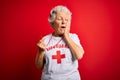 Senior beautiful grey-haired lifeguard woman wearing t-shirt with red cross using whistle Yawning tired covering half face, eye