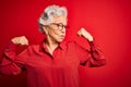 Senior beautiful grey-haired woman wearing casual shirt and glasses over red background showing arms muscles smiling proud
