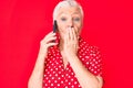 Senior beautiful woman with blue eyes and grey hair having conversation talking on the smartphone covering mouth with hand, Royalty Free Stock Photo