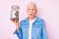 Senior beautiful woman with blue eyes and grey hair holding jar with savings scared and amazed with open mouth for surprise,