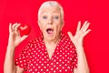 Senior beautiful woman with blue eyes and grey hair holding read heart object scared and amazed with open mouth for surprise,