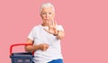 Senior beautiful woman with blue eyes and grey hair holding supermarket shopping basket with open hand doing stop sign with Royalty Free Stock Photo